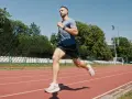 Mężczyzna w sportowym stroju wykonuje szybki marsz na bieżni stadionu, otoczony zielenią i trybunami.