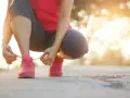 Kobieta w różowej koszulce i szarych legginsach sznuruje różowe buty sportowe. Słońce zachodzi, tworząc ciepłe światło. To idealny moment, by zacząć trening i poczuć szybki marsz efekty.