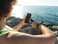Kobieta sprawdza na telefonie, jak połączyć z telefonem swój zegarek fitpro, siedząc nad morzem.