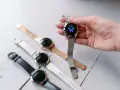 Dłoń trzyma smartwatch z motylem na tarczy. Obok leżą inne zegarki z różnymi paskami: czarnym, białym i różowym.