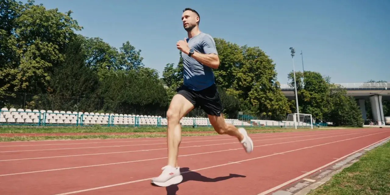 Mężczyzna w sportowym stroju wykonuje szybki marsz na bieżni stadionu, otoczony zielenią i trybunami.