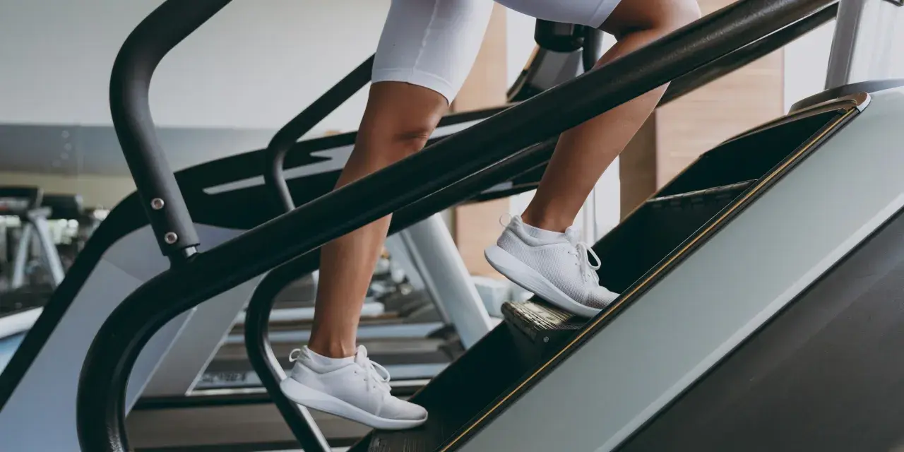 Kobieta w białych spodenkach i butach sportowych wchodzi po schodach na bieżni. To świetny trening cardio, który wzmacnia nogi i poprawia kondycję.