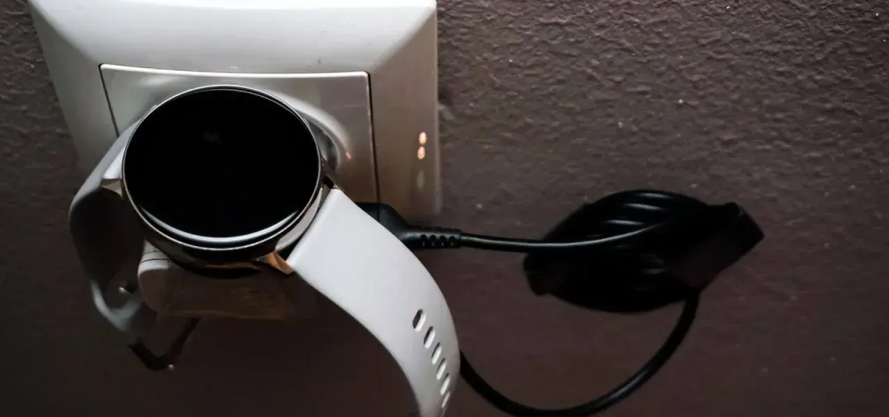 Biały smartwatch z czarnym ekranem podłączony do ładowarki w gniazdku. Prosty sposób, jak ładować smartwatch, by był gotowy do użycia.