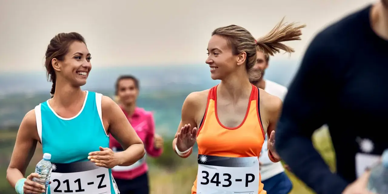 Uśmiechnięte biegaczki na polskim półmaratonie. Dwie kobiety w sportowych strojach biegną, jedna trzyma butelkę wody.