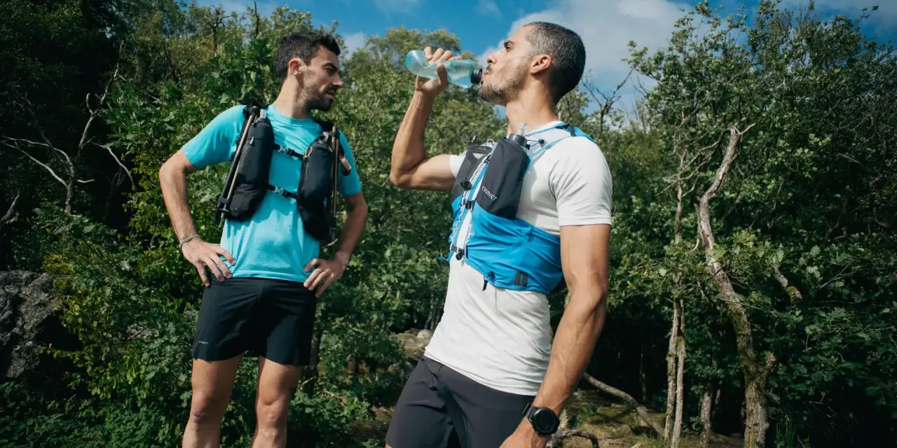 Dwóch biegaczy w kamizelkach nawadniających, jeden pije wodę. Klucz do tego, jak przygotować się do maratonu, to odpowiednie nawodnienie i trening.