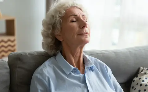 Starsza kobieta z zamkniętymi oczami, oddycha głęboko nosem, relaksując się na kanapie.