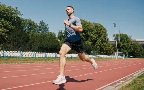Mężczyzna w sportowym stroju wykonuje szybki marsz na bieżni stadionu, otoczony zielenią i trybunami.