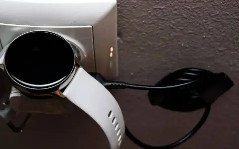 Biały smartwatch z czarnym ekranem podłączony do ładowarki w gniazdku. Prosty sposób, jak ładować smartwatch, by był gotowy do użycia.
