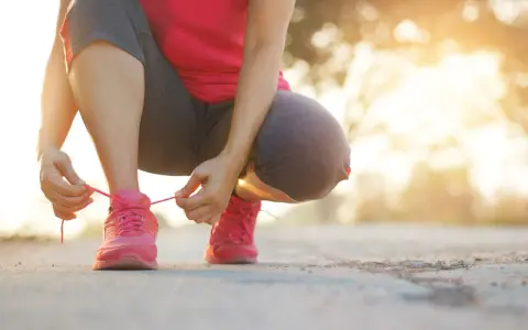 Kobieta w różowej koszulce i szarych legginsach sznuruje różowe buty sportowe. Słońce zachodzi, tworząc ciepłe światło. To idealny moment, by zacząć trening i poczuć szybki marsz efekty.