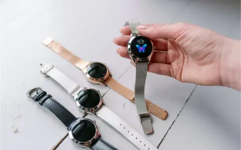 Dłoń trzyma smartwatch z motylem na tarczy. Obok leżą inne zegarki z różnymi paskami: czarnym, białym i różowym.
