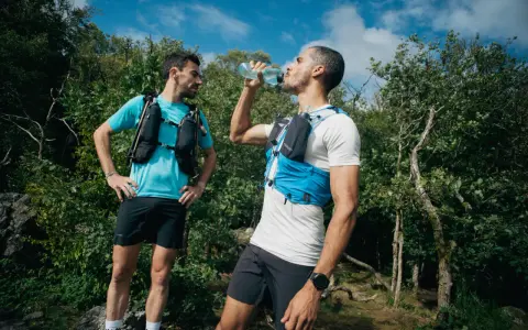 Dwóch biegaczy w kamizelkach nawadniających, jeden pije wodę. Klucz do tego, jak przygotować się do maratonu, to odpowiednie nawodnienie i trening.
