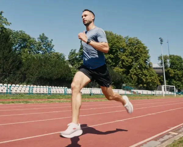Mężczyzna w sportowym stroju wykonuje szybki marsz na bieżni stadionu, otoczony zielenią i trybunami.