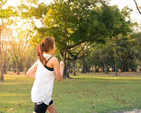 Kobieta w sportowym stroju biegnie przez park. Słońce przebija się przez drzewa, tworząc ciepłą atmosferę. To idealny dzień na realizację jej marszobieg planu.