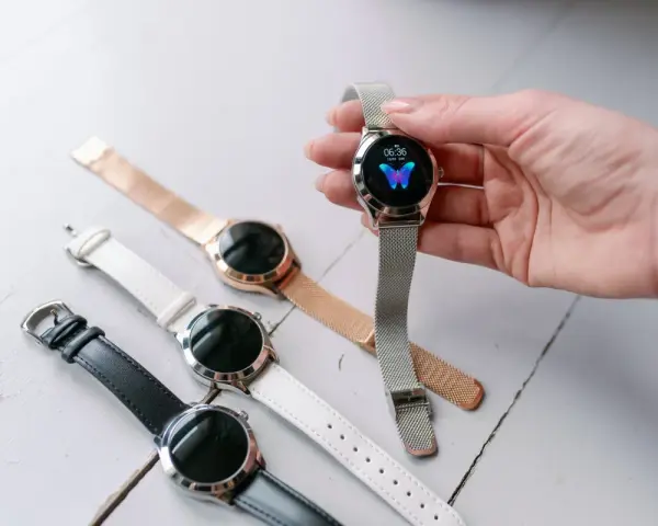 Dłoń trzyma smartwatch z motylem na tarczy. Obok leżą inne zegarki z różnymi paskami: czarnym, białym i różowym.