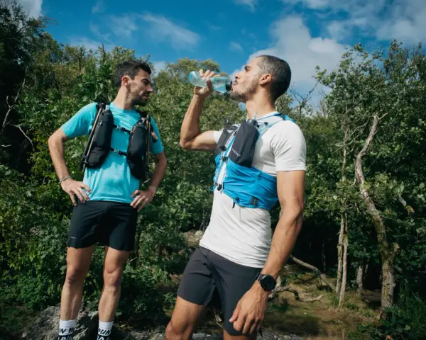 Dwóch biegaczy w kamizelkach nawadniających, jeden pije wodę. Klucz do tego, jak przygotować się do maratonu, to odpowiednie nawodnienie i trening.