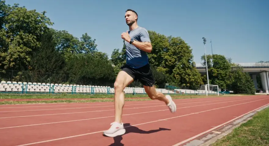 Mężczyzna w sportowym stroju wykonuje szybki marsz na bieżni stadionu, otoczony zielenią i trybunami.