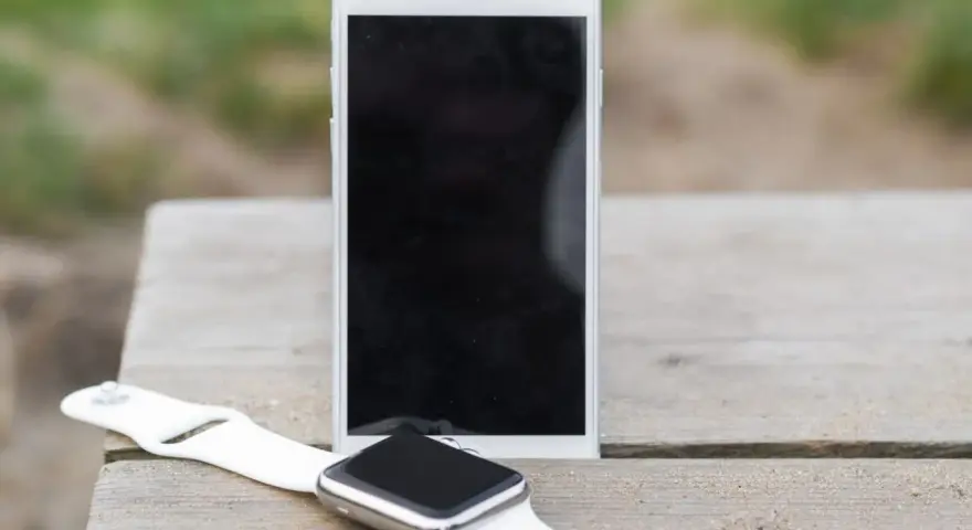 Biały smartwatch i telefon leżą na drewnianym blacie. Dowiedz się, jak zainstalować smartwatch, by połączyć go z telefonem.