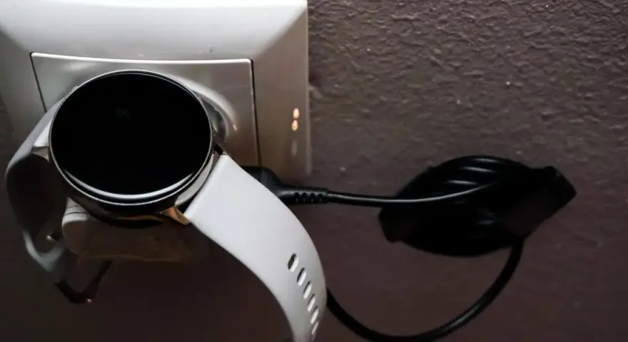 Biały smartwatch z czarnym ekranem podłączony do ładowarki w gniazdku. Prosty sposób, jak ładować smartwatch, by był gotowy do użycia.