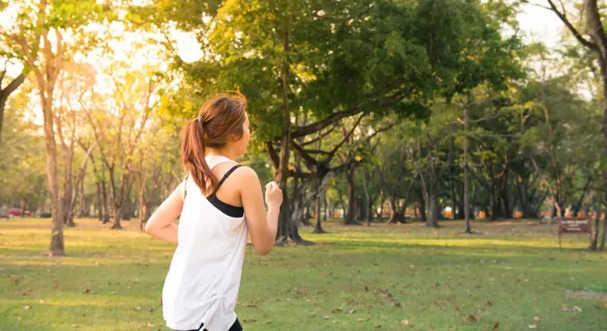 Kobieta w sportowym stroju biegnie przez park. Słońce przebija się przez drzewa, tworząc ciepłą atmosferę. To idealny dzień na realizację jej marszobieg planu.