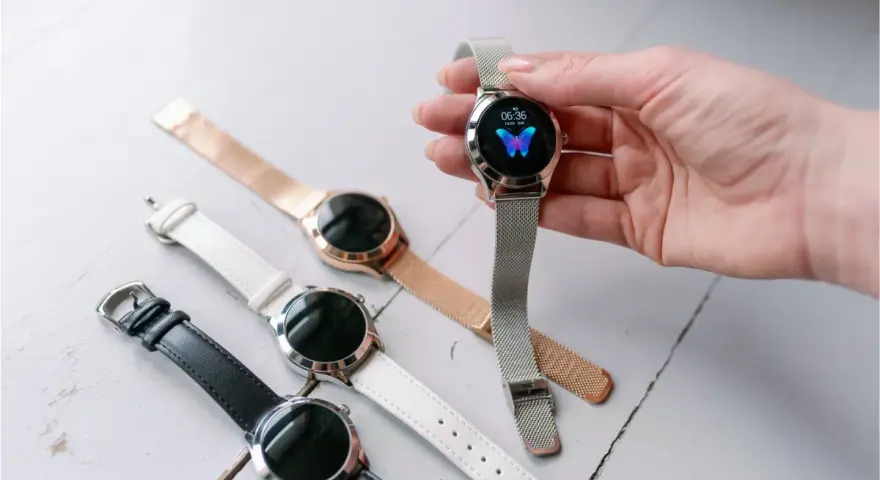 Dłoń trzyma smartwatch z motylem na tarczy. Obok leżą inne zegarki z różnymi paskami: czarnym, białym i różowym.