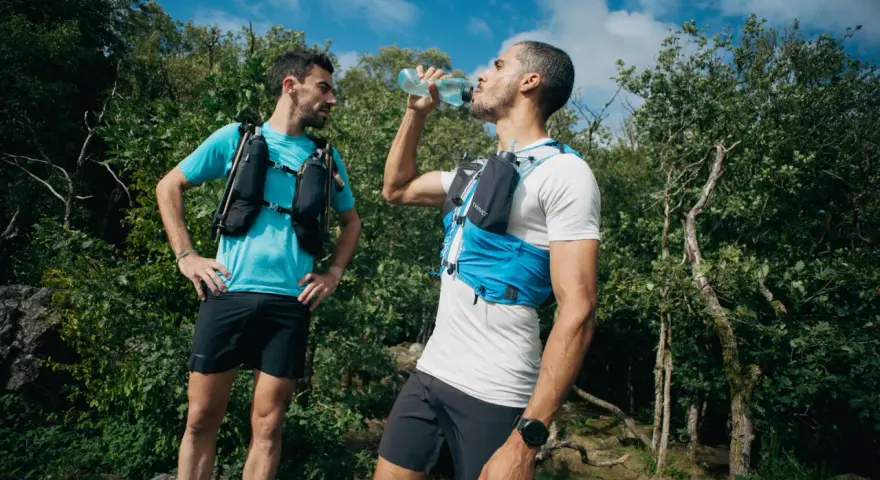 Dwóch biegaczy w kamizelkach nawadniających, jeden pije wodę. Klucz do tego, jak przygotować się do maratonu, to odpowiednie nawodnienie i trening.