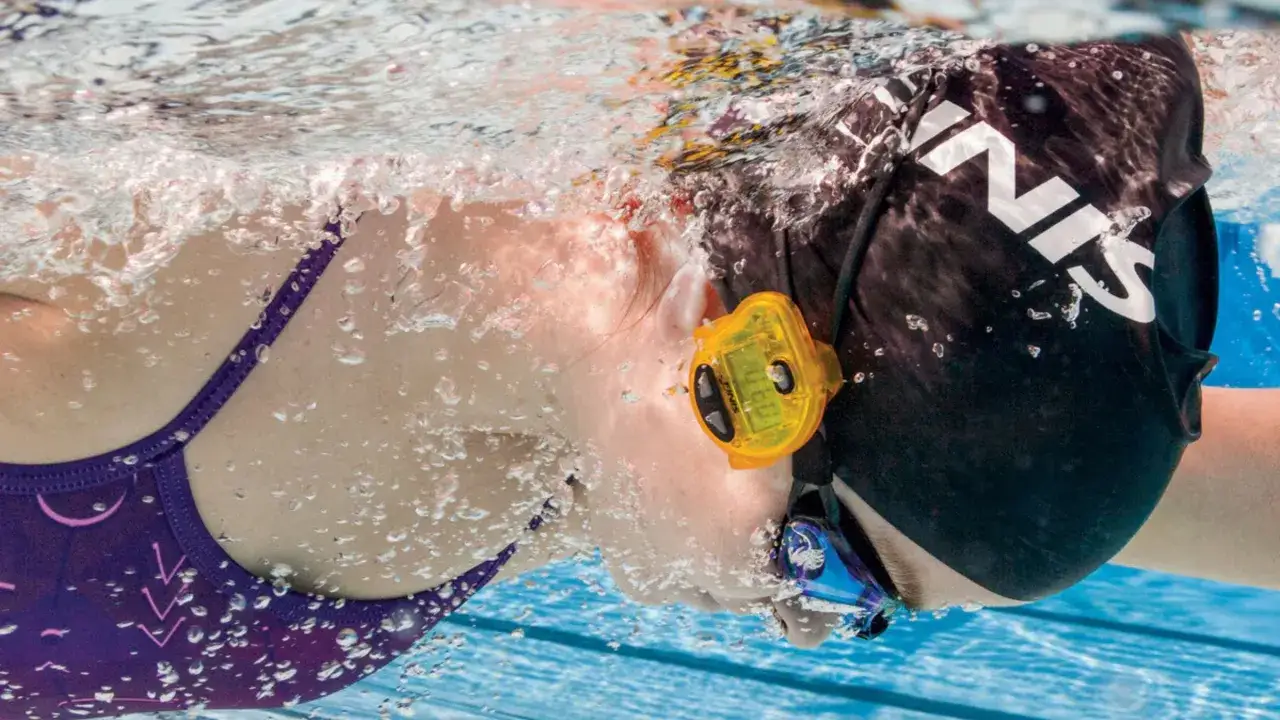Pływaczka w czepku i okularach z żółtym stoperem, mierzy średnie tempo pływania na 100m.