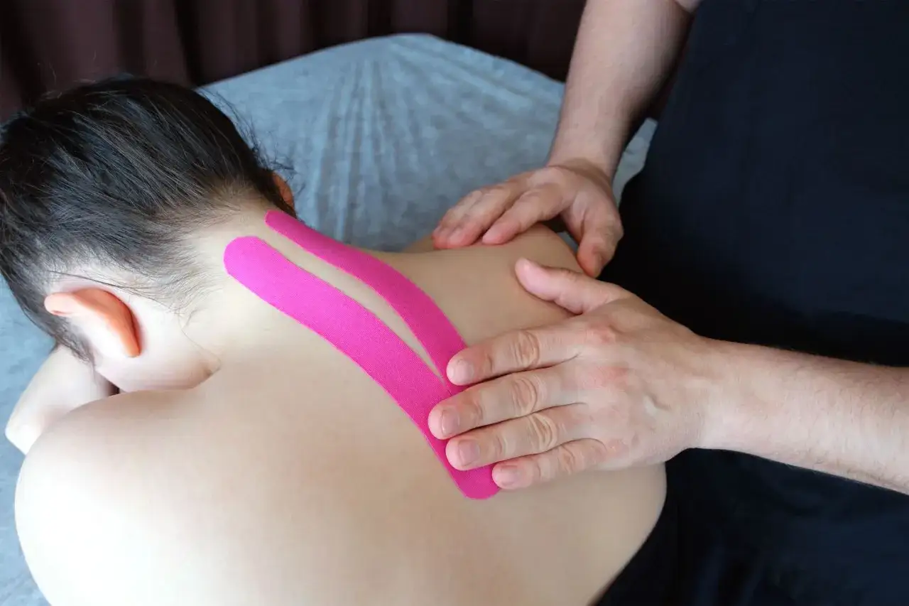 Taping barku i szyi. Terapeuta aplikuje różową taśmę kinezjologiczną na plecy i kark pacjentki, wspierając mięśnie.