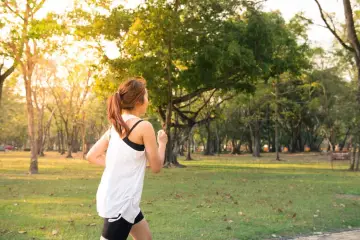 Kobieta w sportowym stroju biegnie przez park. Słońce przebija się przez drzewa, tworząc ciepłą atmosferę. To idealny dzień na realizację jej marszobieg planu.