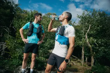 Dwóch biegaczy w kamizelkach nawadniających, jeden pije wodę. Klucz do tego, jak przygotować się do maratonu, to odpowiednie nawodnienie i trening.