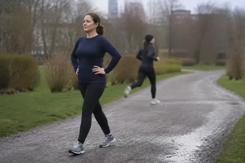 Kobieta w ciemnym stroju sportowym spaceruje po mokrej ścieżce w parku. W tle inna osoba biegnie. 12 km/h jakie to tempo dla niej?